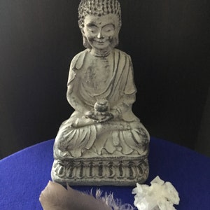 May include: A white stone Buddha statue sitting in meditation pose on a blue surface with a gray feather and a white crystal beside it.