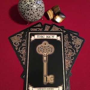May include: A tarot card featuring a gold key design on a black background. The card reads "The Key" and "Locked doors can now be opened". The card is surrounded by other tarot cards and a speckled stone sphere.