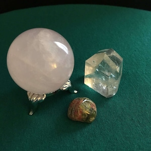 May include: Three polished stones: a large, round, pale pink rose quartz sphere on a silver stand, a clear, faceted quartz crystal, and a smaller, round, green, red, and brown jasper stone.