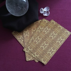 Könnte beinhalten: Vier Tarotkarten mit einem gold-braunen Muster liegen auf einer dunkelvioletten Oberfläche. Eine klare Kristallkugel liegt auf einem schwarzen Tuch und drei klare Quarzkristalle sind in der Nähe verstreut.