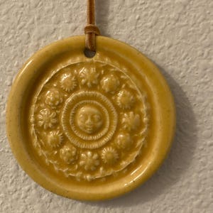 May include: A round, yellow ceramic wall hanging with a detailed floral and face design. The decorative piece is suspended by a brown leather cord. The wall hanging is a decorative item.