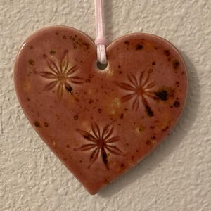 May include: A heart-shaped ceramic ornament in a reddish-brown hue, adorned with three embossed starburst designs. It hangs from a pale pink ribbon, offering a touch of rustic charm. The ornament is likely handmade and adds a decorative element to any space.