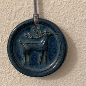 May include: A round, blue ceramic ornament with a raised image of a deer. The deer is depicted in a simple, linear style. The ornament is suspended by a gray ribbon. The background is a textured white wall.