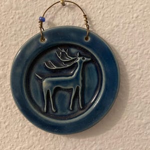May include: Round, blue ceramic wall hanging featuring a deer silhouette. The deer is in a raised relief design. The hanging has a brass wire and a blue bead for hanging. The ceramic is a glossy, dark blue color.