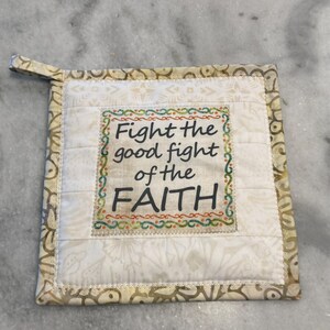May include: Square quilted pot holder with the text "Fight the good fight of the FAITH." The pot holder is cream-colored with a patterned border and a small loop for hanging. The text is in black and green.