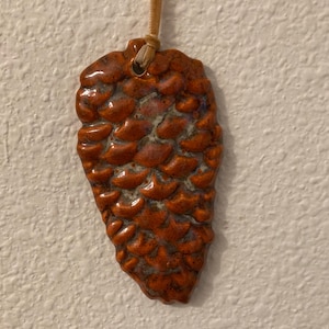 May include: A ceramic pinecone pendant with a textured surface and a warm, reddish-brown glaze. The pendant is suspended from a tan cord. The pinecone design is detailed and realistic, showcasing the natural form.