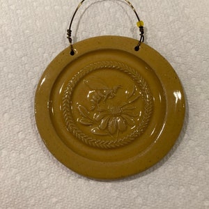 May include: A round, brown ceramic ornament with a raised design of a bee and flower. The ornament has a wire hanger with a yellow bead.