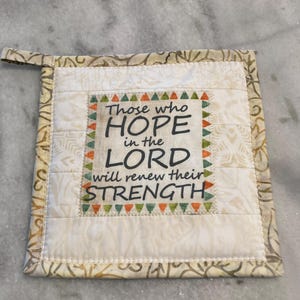 May include: Square quilted pot holder with the text "Those who hope in the Lord will renew their strength." The pot holder is cream-colored with a patterned border and a loop for hanging.