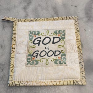 May include: Square quilted pot holder with the words "GOD is GOOD" in black script. The center panel is cream-colored with a floral design, framed by a decorative border. A small loop is attached for hanging.