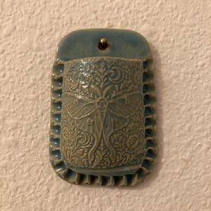 May include: A blue-grey ceramic wall pocket with a flat back and a rounded, ruffled edge. The front surface features an embossed design of a large moth centered among intricate floral and vine patterns, with a small hole at the top for hanging.