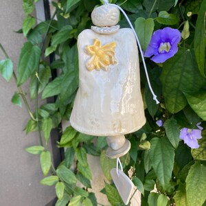 May include: A cream-colored ceramic wind chime with a yellow bee design. The bell-shaped chime hangs from a white cord, surrounded by green leaves and purple flowers. The chime has a textured surface and a small, heart-shaped tag.
