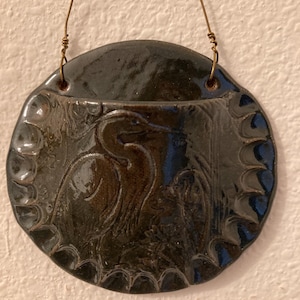 May include: A dark, round ceramic wall hanging with a bird relief design. The bird is depicted with long neck and beak. The hanging has a scalloped edge and is suspended by a thin wire. The ceramic is glazed in a dark, glossy finish.