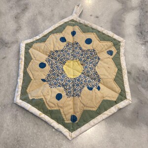 May include: A quilted, hexagonal hot pad with a floral design. The center features a yellow flower surrounded by blue and white patterned fabric. Cream-colored hexagons are outlined in green, with blue dots. A white loop is attached for hanging.