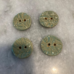 May include: Four round ceramic buttons featuring a glossy teal glaze and a textured leaf pattern. Each button has two central holes for attachment. The surface of the buttons shows a subtle variation in color, blending light green and blue tones.