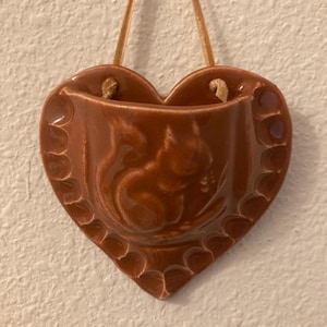 May include: A heart-shaped, brown ceramic wall pocket with a squirrel design. The pocket has a scalloped edge and is suspended by a tan cord. This decorative piece is suitable for small plants or other items.