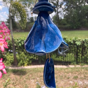 May include: A hanging ceramic wind chime in a deep blue glaze. The chime has a bell-like shape with a layered top and a teardrop-shaped bottom piece. It hangs from a gold-colored cord, with a blurred background of greenery and sky.