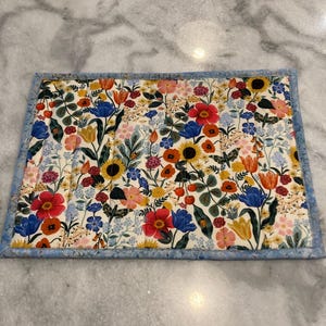 May include: Rectangular floral placemat featuring a vibrant array of flowers in red, blue, yellow, and pink on a cream background. The placemat has a light blue border and is likely made of fabric.