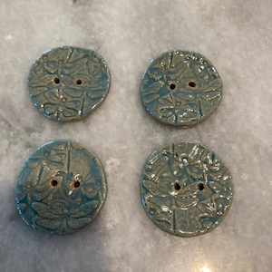 May include: Four round ceramic buttons in a blue-green glaze, each featuring a debossed dragonfly pattern and two central holes for attachment. The buttons have a slightly irregular, organic circular shape with a glossy, textured surface finish.