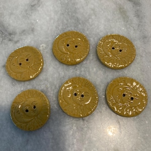 May include: Six circular mustard yellow buttons, each featuring a raised relief design of a sun and crescent moon face. The buttons have a glossy finish and two central holes for attachment, arranged in a scattered pattern on a light surface.