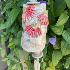 May include: A handmade ceramic wind chime with a cylindrical body and a textured, off-white glaze. The chime features three dimensional red and yellow flower accents. The chime hangs from a white string, surrounded by green foliage.