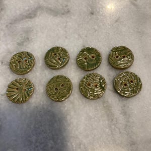 May include: Eight round, olive green ceramic buttons arranged in two rows. Each button features a glossy, textured surface with an embossed botanical leaf pattern and two small holes in the center for attachment to fabric or other materials.