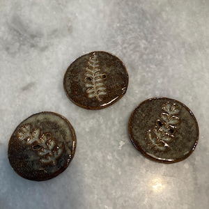 May include: Three circular ceramic buttons featuring a dark brown, textured glaze with a glossy finish. Each button displays a central, light-colored fern leaf imprint with visible veins, and two small holes for attachment in the center of the leaf.