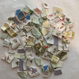 May include: A collection of assorted ceramic shards featuring various floral patterns, solid pastel colors, and gold-trimmed edges. These broken pieces of porcelain display intricate botanical illustrations, including roses and leaves, in a mix of white, blue, green, and purple tones.