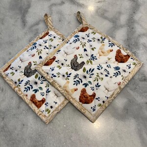 May include: Two square quilted pot holders with a white background featuring a repeating pattern of chickens, blue leaves, and white flowers. The edges are trimmed with a tan and cream patterned fabric, and each has a matching loop for hanging.