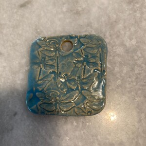 May include: A square ceramic pendant with rounded corners, featuring a glossy light blue glaze and an embossed pattern of multiple dragonflies. The piece has a single circular hole at the top center for hanging, with a smooth, reflective finish.