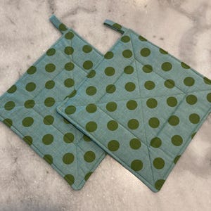 May include: Two square potholders with a light blue background and large olive green polka dots. Each potholder has a small loop for hanging. The potholders are quilted with diagonal stitching.