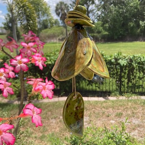 May include: A handcrafted ceramic wind chime with a unique, layered design. The wind chime features a green and brown glaze, with a teardrop-shaped pendant hanging below. The chime is suspended by a natural-colored cord, perfect for outdoor decor.