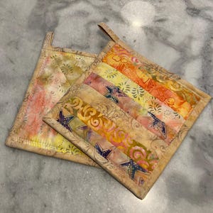 May include: Two quilted pot holders with a colorful, patchwork design. One pot holder features a mix of orange, yellow, and pink stripes with star accents. The other has a floral pattern in similar colors. Both have a loop for hanging.