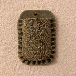 May include: A rectangular wall-mounted ceramic pocket with a sage green glaze. The front surface features a relief design of birds perched among intricate branches and leaves. The edges have a ruffled texture, and a single hole is at the top.