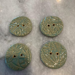 May include: Four round, sage green ceramic buttons, each featuring a detailed, embossed tropical leaf pattern across the surface. Every button has two small, centered holes for attachment, with a glossy, smooth finish on the textured clay.