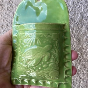 May include: A lime green ceramic wall pocket planter featuring a raised relief design of a sea turtle swimming among coral and seashells. The rectangular vessel has a scalloped border and a rounded top edge with a hole for hanging on a wall.