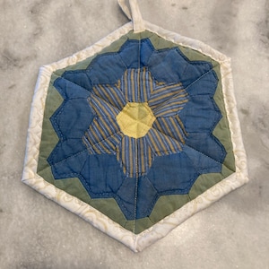 May include: Hexagon-shaped quilted potholder with a floral design. The center features a yellow and blue striped pattern, surrounded by blue and green geometric shapes. The edges are finished with a white patterned fabric and a hanging loop.