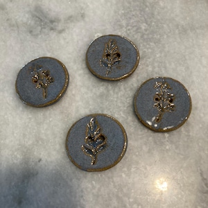 May include: Four circular ceramic buttons with a speckled blue-grey glaze and a brown rim. Each button features a debossed botanical leaf or sprig design in the center. The buttons have two holes for attachment and a smooth, glossy finish.