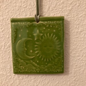 May include: Green ceramic wall hanging with celestial design. The square plaque features a crescent moon, sun with a smiling face, stars, and decorative border. A green ribbon is attached for hanging.