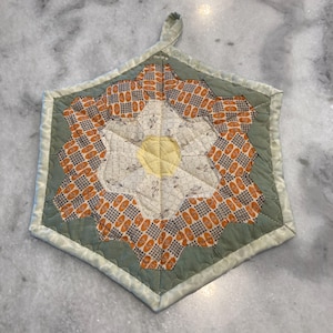 May include: A quilted, hexagonal hot pad with a loop for hanging. The center features a yellow circle surrounded by white and orange patterned fabric. The outer edge is a muted green with a light green trim.