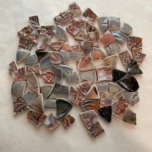 May include: A collection of assorted ceramic mosaic tiles in various shapes and sizes. The pieces feature a mix of colors including dusty pink, soft blue, grey, and black, with some tiles displaying intricate embossed floral and scroll patterns.