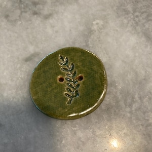 May include: A round, olive green ceramic button featuring a glossy finish and a debossed leaf sprig design in the center. The button has two small holes positioned on either side of the stem of the leaf motif for attachment purposes.