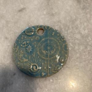 May include: A circular blue ceramic pendant featuring a glossy finish and embossed celestial designs, including a central compass rose, stars, and crescent moons. The piece has a single hole at the top for hanging, with a smooth, glazed surface.