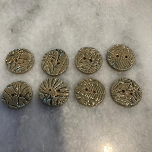 May include: Eight round, tan-colored buttons arranged in two rows of four. Each button features a textured, embossed leaf pattern across its surface and contains two small holes in the center for attachment to fabric or other materials.