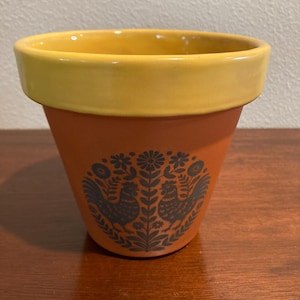 May include: A terracotta plant pot with a wide, smooth yellow rim. The exterior features a dark gray circular graphic print depicting two roosters facing each other, surrounded by stylized floral and leaf patterns on the orange surface.