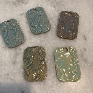 May include: Five rectangular ceramic pendants with rounded corners and a single hole at the top. Each piece features a raised relief design of mermaids in various glaze colors including light blue, dark blue, olive green, and seafoam green.