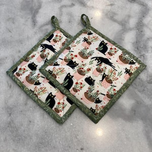 May include: Two square pot holders with a white background featuring a repeating pattern of black cats, pink potted plants, and red flowers. The edges are trimmed with a green leaf pattern and have a green loop for hanging.