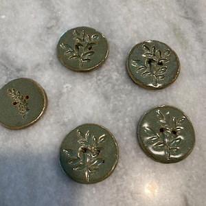 May include: Five circular ceramic buttons in a sage green glaze, each featuring a debossed botanical leaf pattern. The buttons have a glossy finish with visible brown edges and two small holes in the center for attachment to garments or crafts.