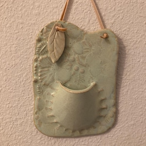 May include: A light green ceramic wall pocket with a leaf accent and a tan leather hanging strap. The pocket has a textured surface with floral and dot patterns, and a small, curved opening.