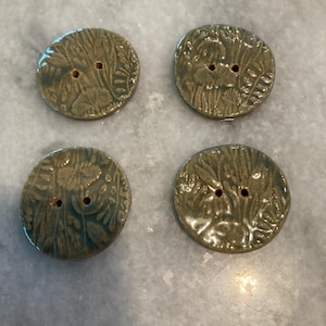 May include: Four round ceramic buttons with a glossy sage green glaze. Each button features a two-hole design and an embossed botanical pattern of leaves and flowers across the surface. The edges of the buttons are slightly irregular and rounded.