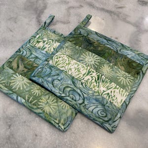 May include: Two square quilted potholders featuring a patchwork design of batik fabric in shades of blue, sage green, and white. The fabric displays patterns of swirling lines, floral motifs, and starbursts, with each potholder including a small fabric loop.